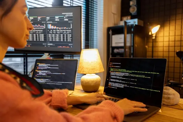 A developer working on multiple laptops with coding on display and data analytics on a screen, representing project outsourcing in a modern tech environment.